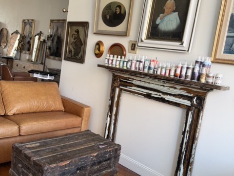 A salvaged, paint-worn fireplace mantel lined with color cans, under a hung group of gilt-framed portrait paintings, beside a leather sofa and an antique steamer trunk.