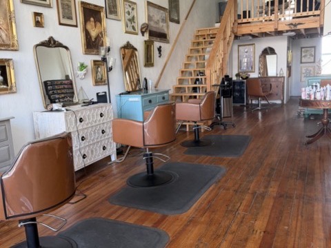 Styling chairs on mats down a row of antique dressers used as stations, beneath a wall of gilt-framed pictures, with the open timber stair and loft behind.