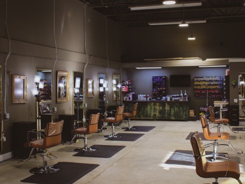 The main salon floor, styling chairs on mats down a wall of gilt mirrors and ring lights, with the color bar and shelves of tube color across the back wall.