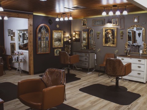A styling room at Craft Collective with a wood-plank ceiling and warm lighting, its walls hung with gilt-framed paintings, antique mirrors and stained-glass panels above tan leather chairs at vintage dresser stations.